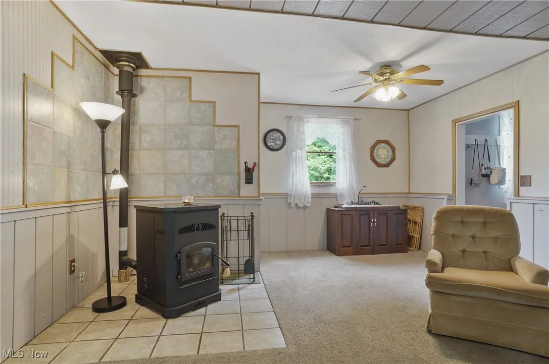 Living area with a decorative wall, a wood stove, carpet, wainscoting, and ceiling fan