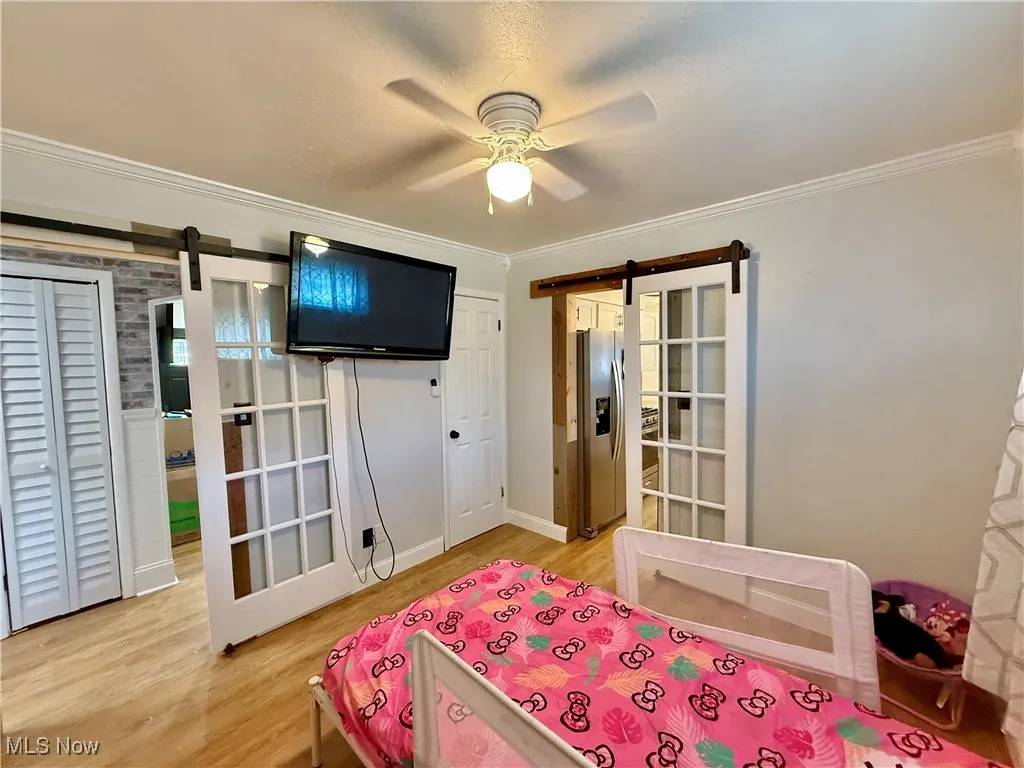 Bedroom with a barn door, crown molding, light wood-type flooring, stainless steel fridge, and ceiling fan