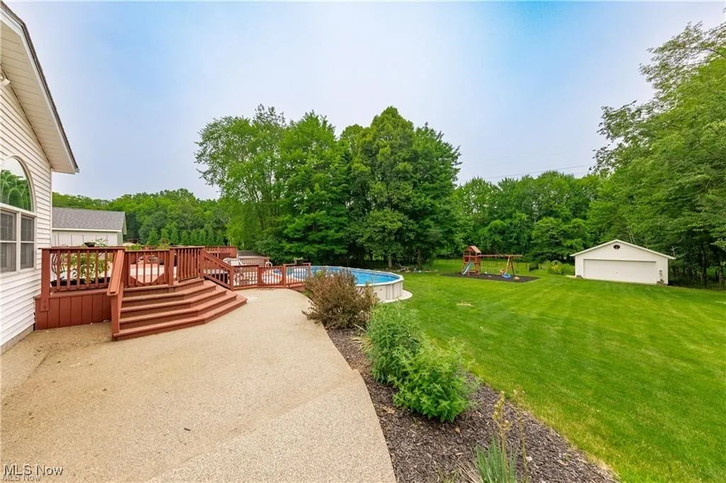 View of green lawn with an outdoor pool, a playground, an outbuilding, a wooden deck, and a patio area