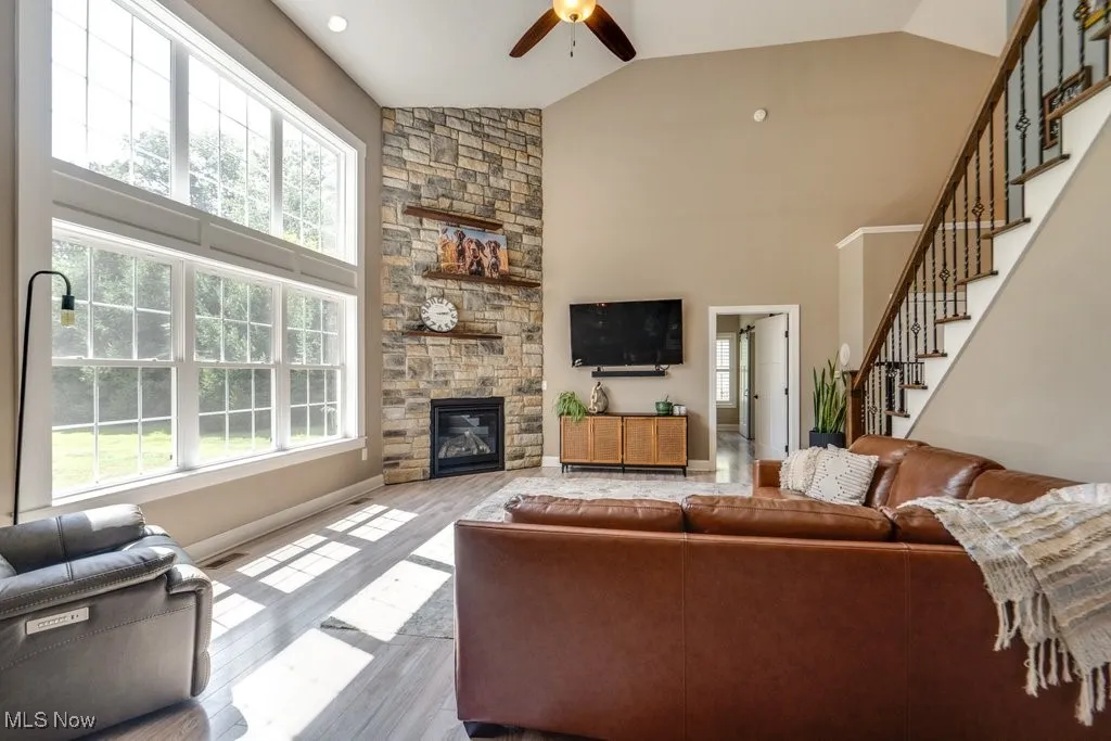 Living room with wood finished floors, a ceiling fan, a stone fireplace, high vaulted ceiling, and stairs