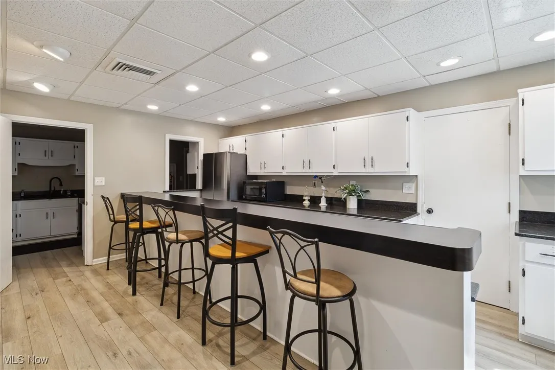 Kitchen with a drop ceiling, a kitchen bar, dark countertops, white cabinets, and light wood-style floors