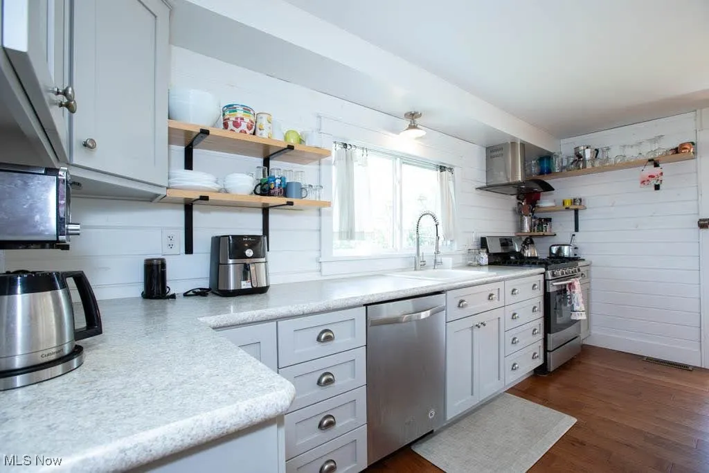 Kitchen with open shelves, stainless steel appliances, light countertops, dark wood-style floors, and wall chimney exhaust hood