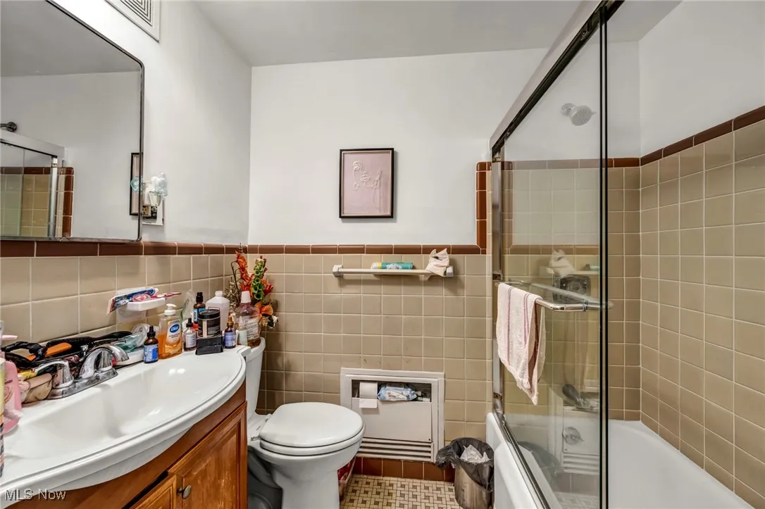 Full bath with shower / bath combination with glass door, vanity, tile walls, heating unit, and a wainscoted wall