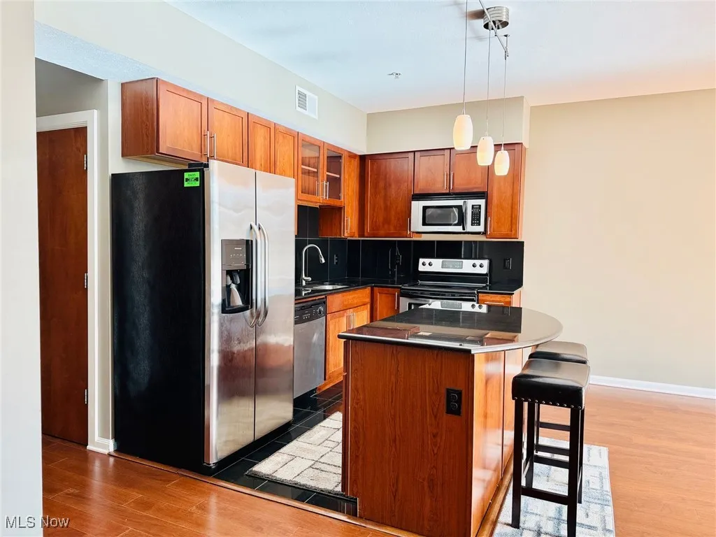 Kitchen featuring stainless steel appliances, glass insert cabinets, brown cabinets, a kitchen bar, and dark wood-style flooring