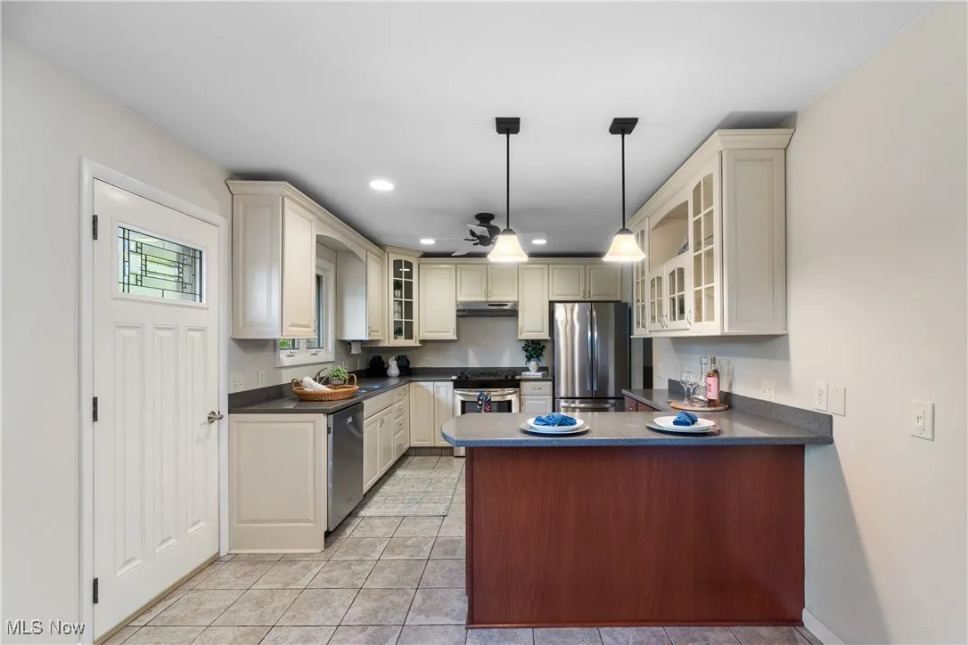 Kitchen with glass insert cabinets, cream cabinetry, a peninsula, appliances with stainless steel finishes, and recessed lighting