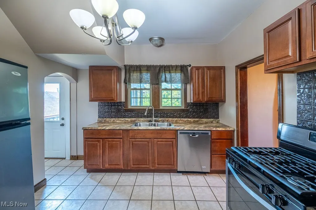 Kitchen with tasteful backsplash, appliances with stainless steel finishes, brown cabinetry, arched walkways, and dark stone countertops