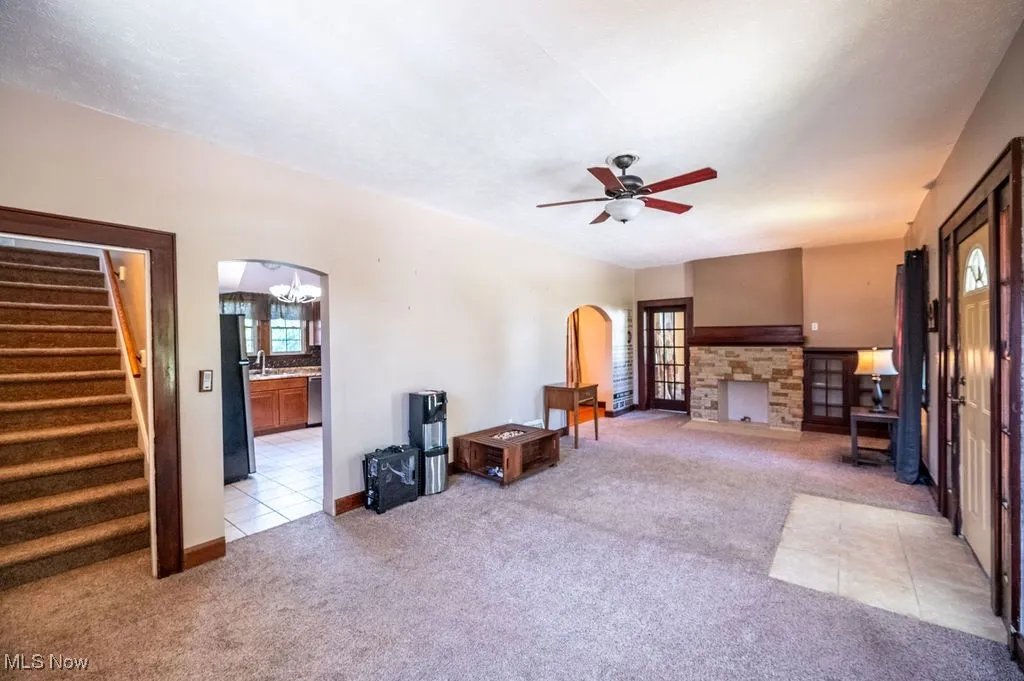 Carpeted living area with stairway, arched walkways, ceiling fan, a stone fireplace, and tile patterned flooring