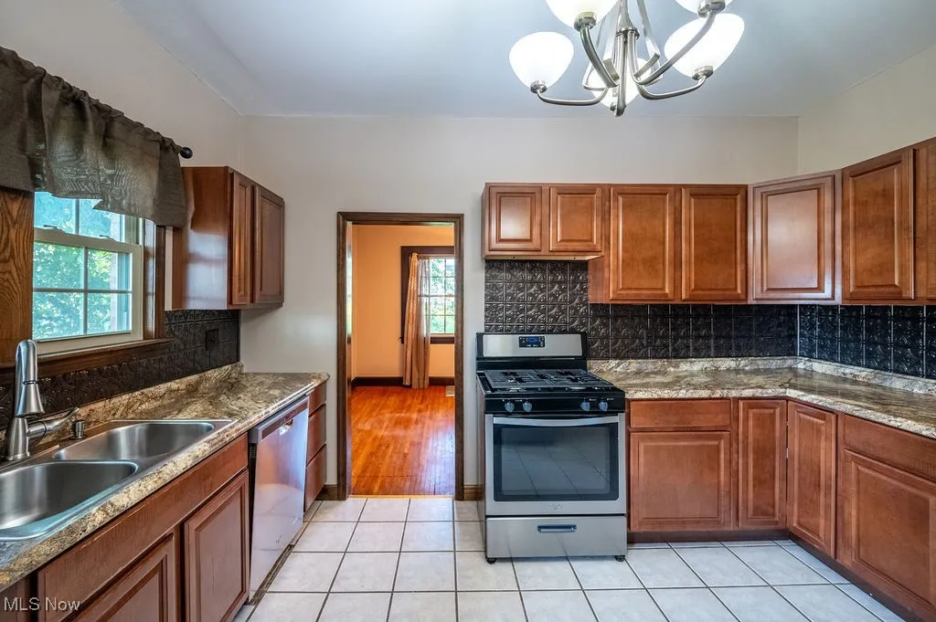 Kitchen featuring decorative backsplash, stainless steel appliances, brown cabinetry, light tile patterned floors, and decorative light fixtures