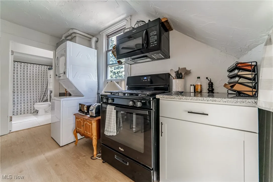 Kitchen with black appliances, stacked washing machine and dryer, light wood-style flooring, white cabinets, and light stone counters
