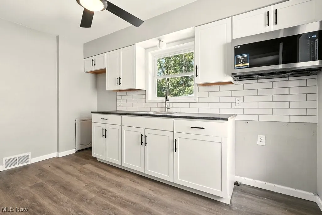 Kitchen featuring dark countertops, stainless steel microwave, white cabinets, dark wood-type flooring, and backsplash