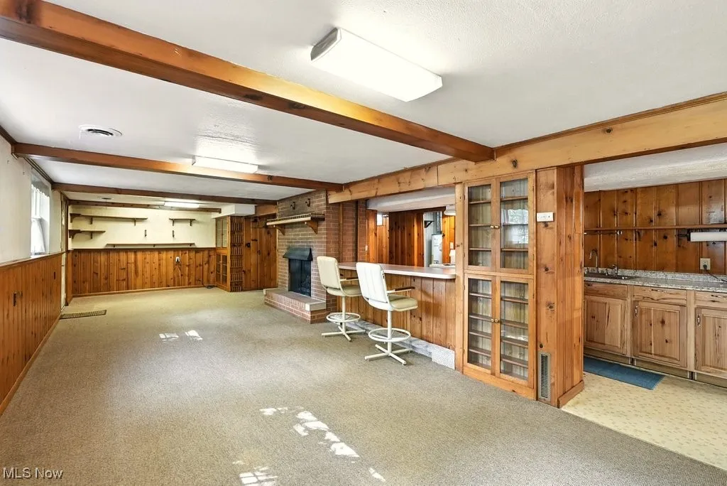 Indoor bar featuring wooden walls, brown cabinetry, light colored carpet, a fireplace, and beam ceiling