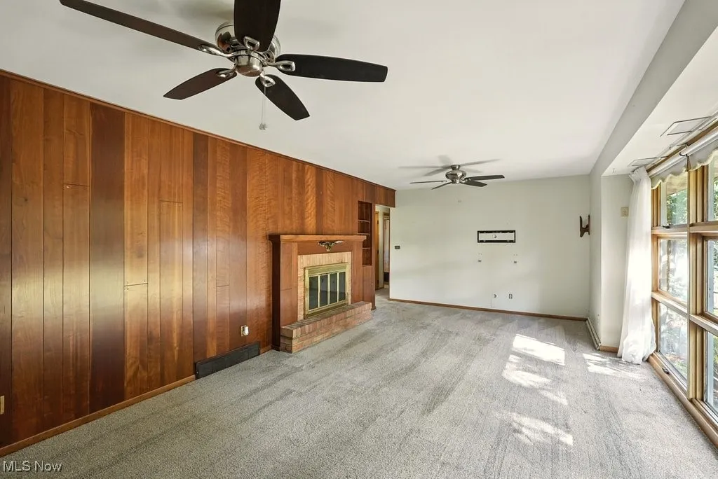 Unfurnished living room with a brick fireplace, carpet flooring, wood walls, and a ceiling fan