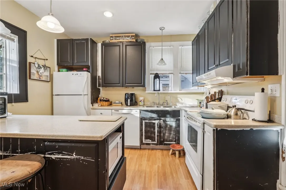 Kitchen with white appliances, hanging light fixtures, light wood-style flooring, under cabinet range hood, and healthy amount of natural light