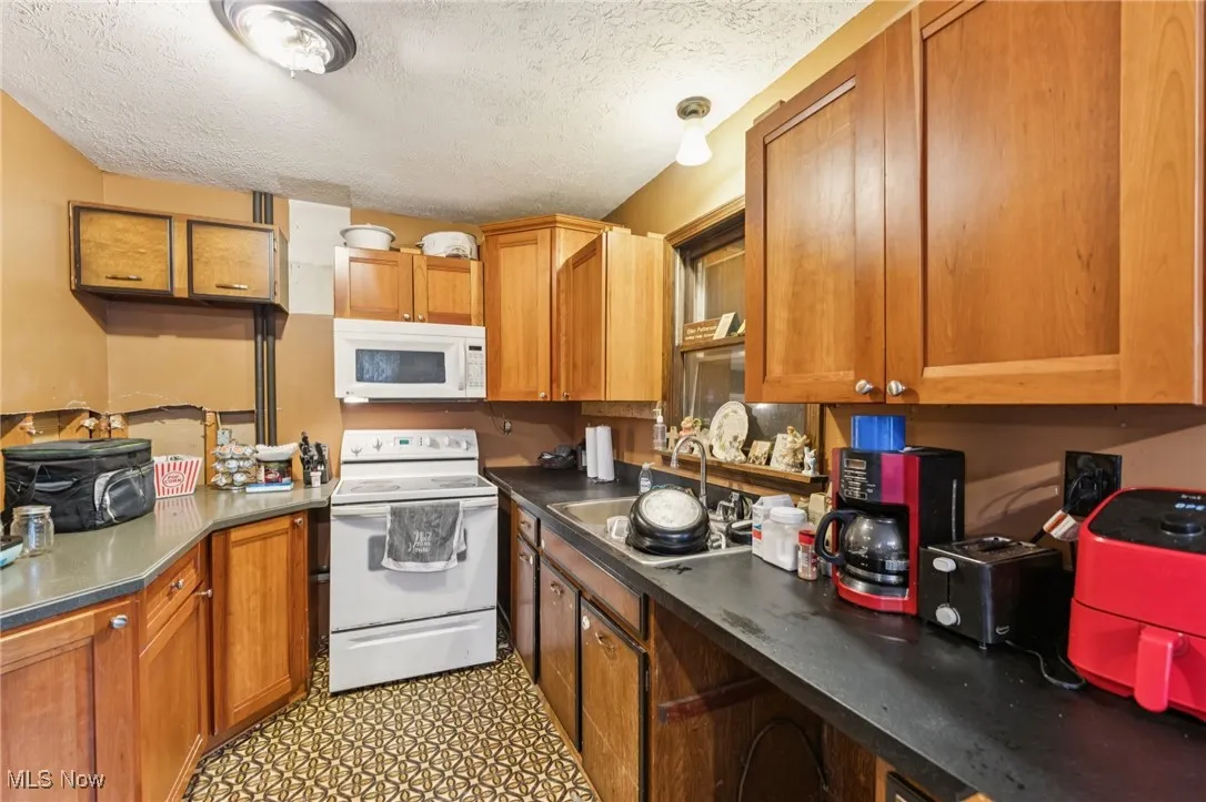 Kitchen featuring white appliances, brown cabinetry, a textured ceiling, and dark countertops