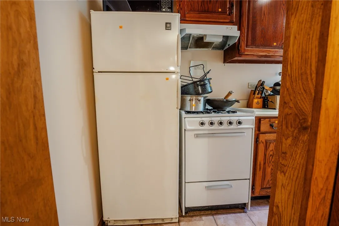 Kitchen featuring white appliances, under cabinet range hood, brown cabinetry, and light tile patterned floors