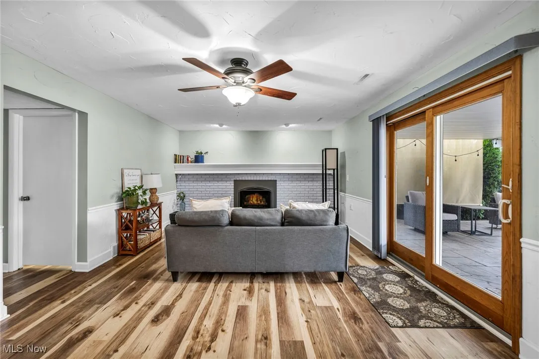 Living room featuring a brick fireplace, wood finished floors, a ceiling fan, and a wainscoted wall