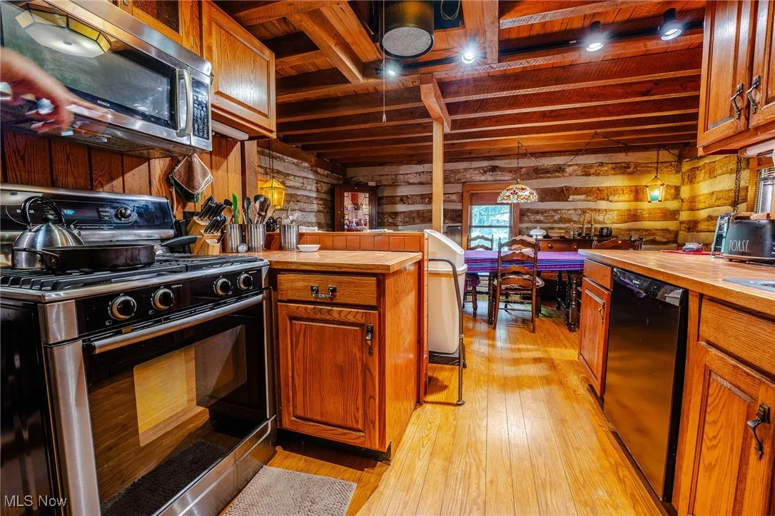 Kitchen featuring range with gas stovetop, stainless steel microwave, wooden walls, brown cabinets, and a wooden ceiling with exposed beams