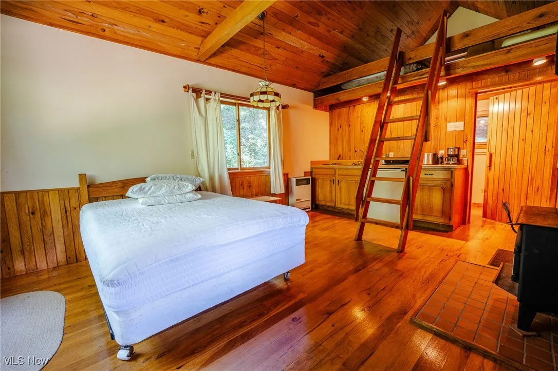 Bedroom with wooden walls, a wood stove, hardwood / wood-style floors, wooden ceiling, and a wainscoted wall