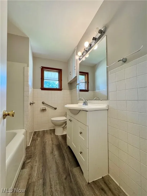Full bath featuring tile walls, dark wood-style flooring, wainscoting, vanity, and a bath