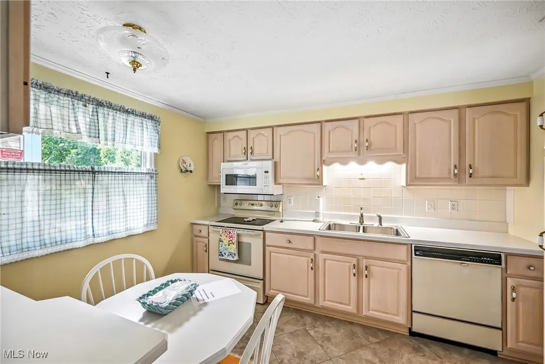 Kitchen with light brown cabinetry, white appliances, backsplash, light countertops, and a textured ceiling