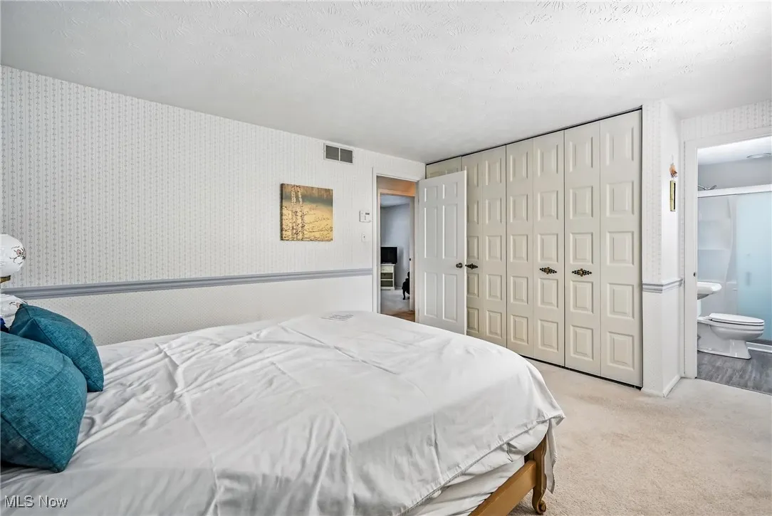 Bedroom with wallpapered walls, light colored carpet, a textured ceiling, a closet, and ensuite bath