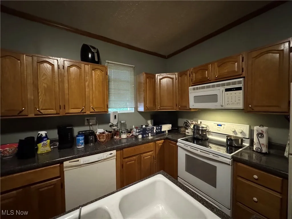 Kitchen featuring white appliances, crown molding, brown cabinetry, a textured ceiling, and dark stone counters