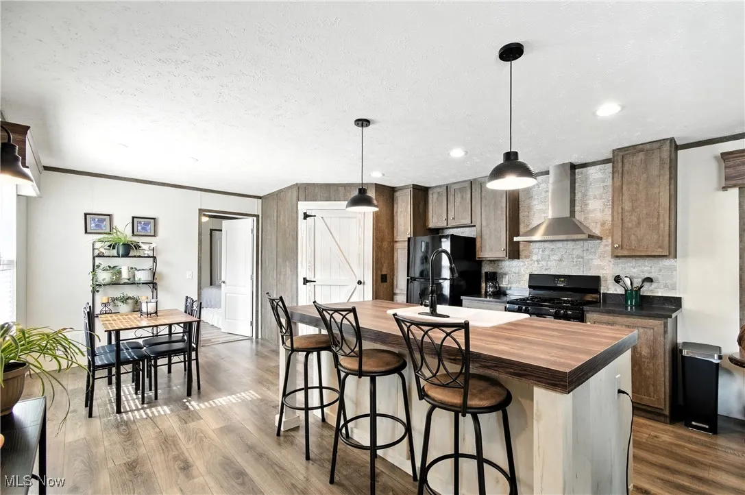 Kitchen with crown molding, a kitchen island with sink, wall chimney exhaust hood, hanging light fixtures, and a breakfast bar
