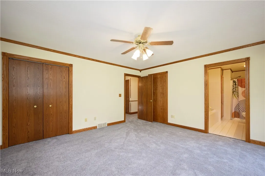 Unfurnished bedroom featuring crown molding, a ceiling fan, ensuite bathroom, carpet, and multiple closets