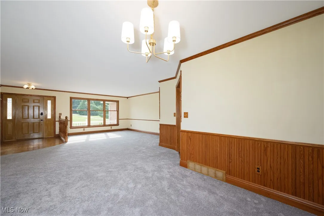 Unfurnished living room with crown molding, a chandelier, carpet, wooden walls, and a wainscoted wall