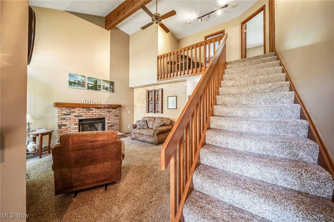 Carpeted living area featuring a brick fireplace, beam ceiling, stairs, high vaulted ceiling, and a ceiling fan