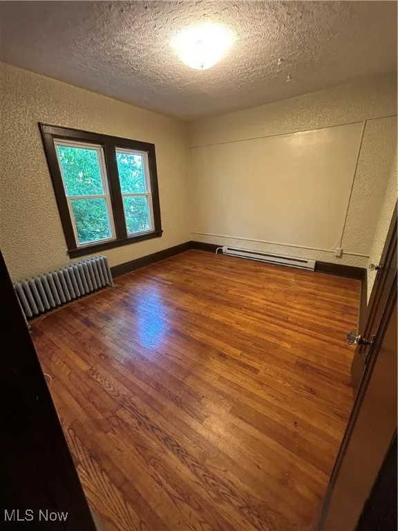 Unfurnished room featuring a textured wall, wood-type flooring, radiator heating unit, baseboard heating, and a textured ceiling