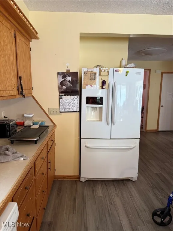 Kitchen with white refrigerator with ice dispenser, dark wood-style floors, light countertops, and brown cabinetry