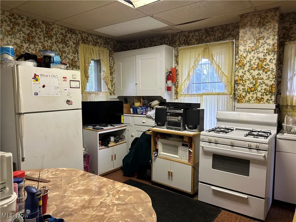 Kitchen with wallpapered walls, a drop ceiling, white cabinets, white appliances, and light countertops
