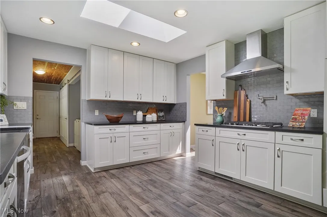 Kitchen featuring a skylight, wall chimney exhaust hood, tasteful backsplash, white cabinets, and dark wood finished floors