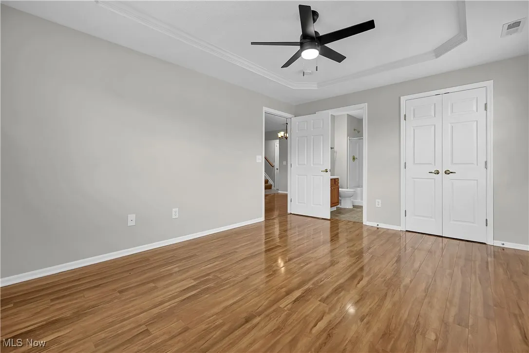 Unfurnished bedroom with a raised ceiling, light wood-style flooring, ceiling fan, connected bathroom, and a closet