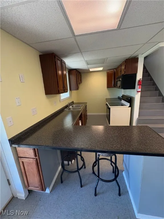 Kitchen with a breakfast bar area, black appliances, dark countertops, a paneled ceiling, and a peninsula