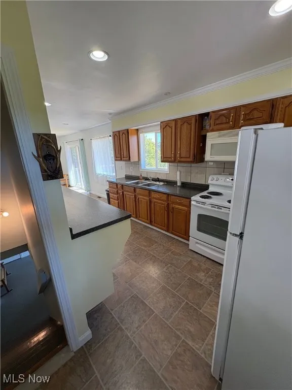 Kitchen featuring brown cabinetry, white appliances, dark countertops, recessed lighting, and tasteful backsplash