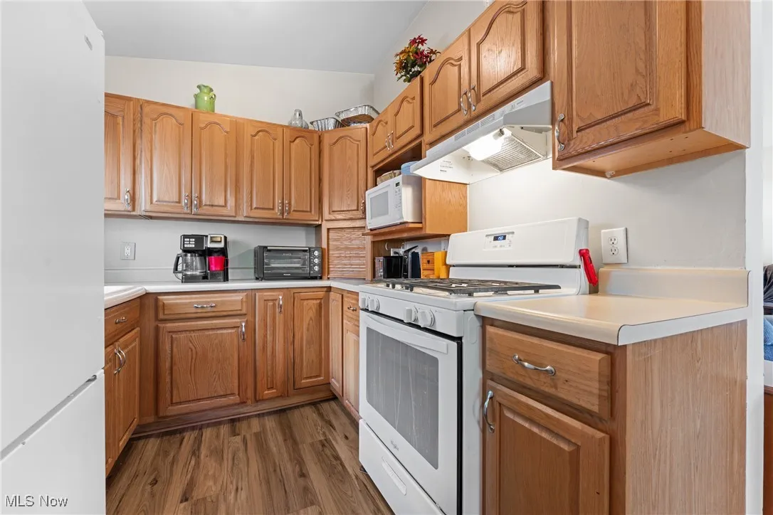 Kitchen with white appliances, light countertops, dark wood finished floors, under cabinet range hood, and brown cabinetry