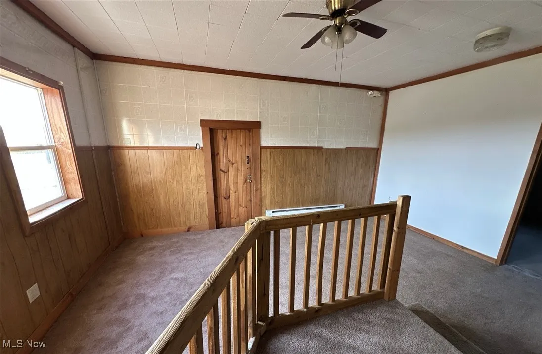 Unfurnished room featuring wooden walls, carpet floors, crown molding, ceiling fan, and a baseboard radiator