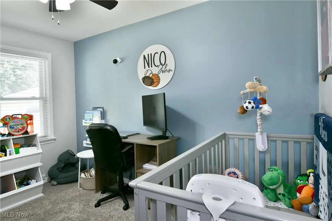 Carpeted bedroom featuring a crib, ceiling fan, and an office area