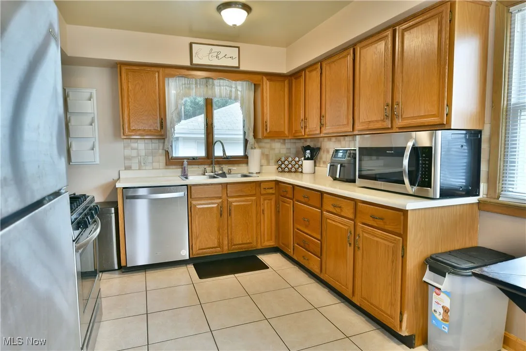 Kitchen with stainless steel appliances, light countertops, backsplash, brown cabinetry, and light tile patterned floors