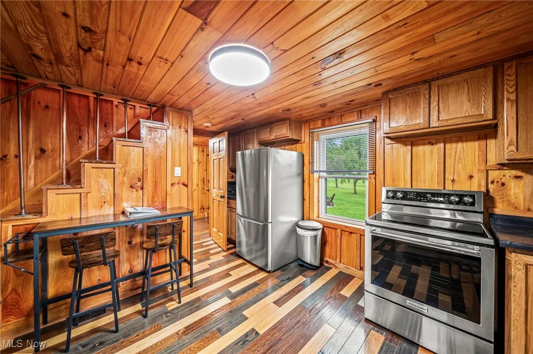 Kitchen with brown cabinetry, wood walls, appliances with stainless steel finishes, wood ceiling, and dark wood-style flooring