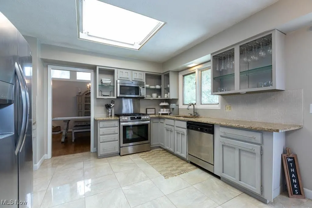 Kitchen featuring gray cabinets, glass insert cabinets, stainless steel appliances, light stone countertops, and open shelves