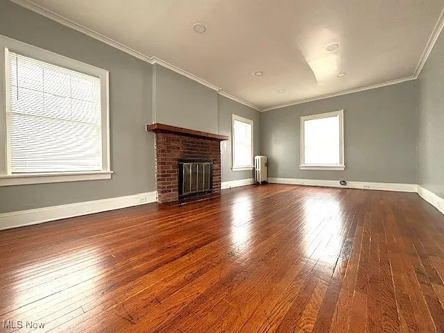 Unfurnished living room with a brick fireplace, dark wood-style flooring, ornamental molding, and radiator