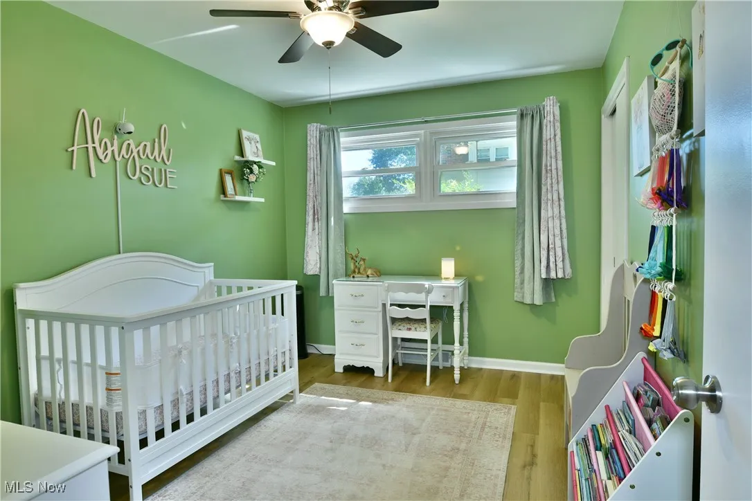 Bedroom with wood finished floors, a crib, a ceiling fan, and a desk