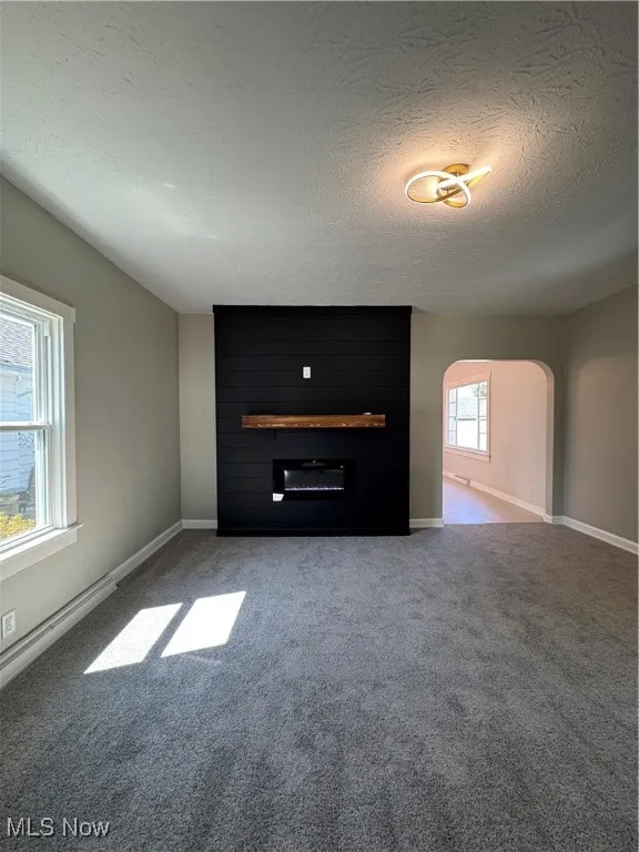 Unfurnished living room with a textured ceiling, light carpet, baseboard heating, arched walkways, and a fireplace