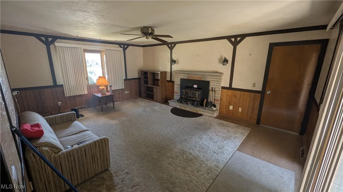 Living room featuring a wood stove, ceiling fan, wood walls, a wainscoted wall, and a textured ceiling