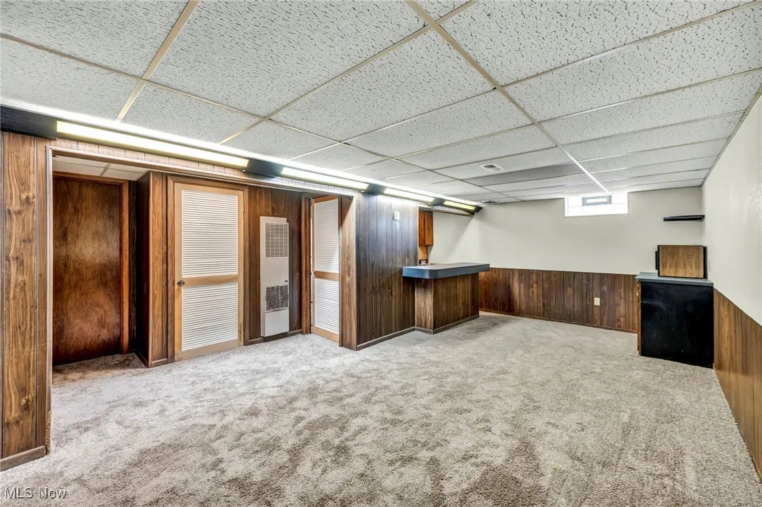 Below grade area with wooden walls, light carpet, a drop ceiling, a heating unit, and a wainscoted wall