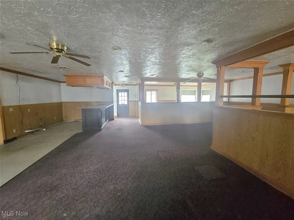 Unfurnished room featuring wood walls, wainscoting, ceiling fan, dark carpet, and a textured ceiling
