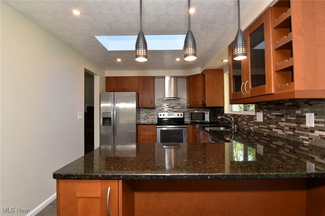 Kitchen featuring brown cabinets, a skylight, dark stone countertops, stainless steel appliances, and wall chimney exhaust hood